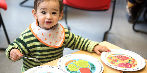 Kleinkind spielt mit drei Kinderteller aus der Mensa und sitzt an einem Tisch.